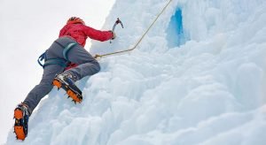 This is a picture of an Alpinist woman with ice tools axe in orange helmet climbing a large wall of ice for an article called Which industries use Competitive Intelligence and why? By Octopus Competitive Intelligence company. We answer the questions with Competitive Intelligence, Due Diligence and business development